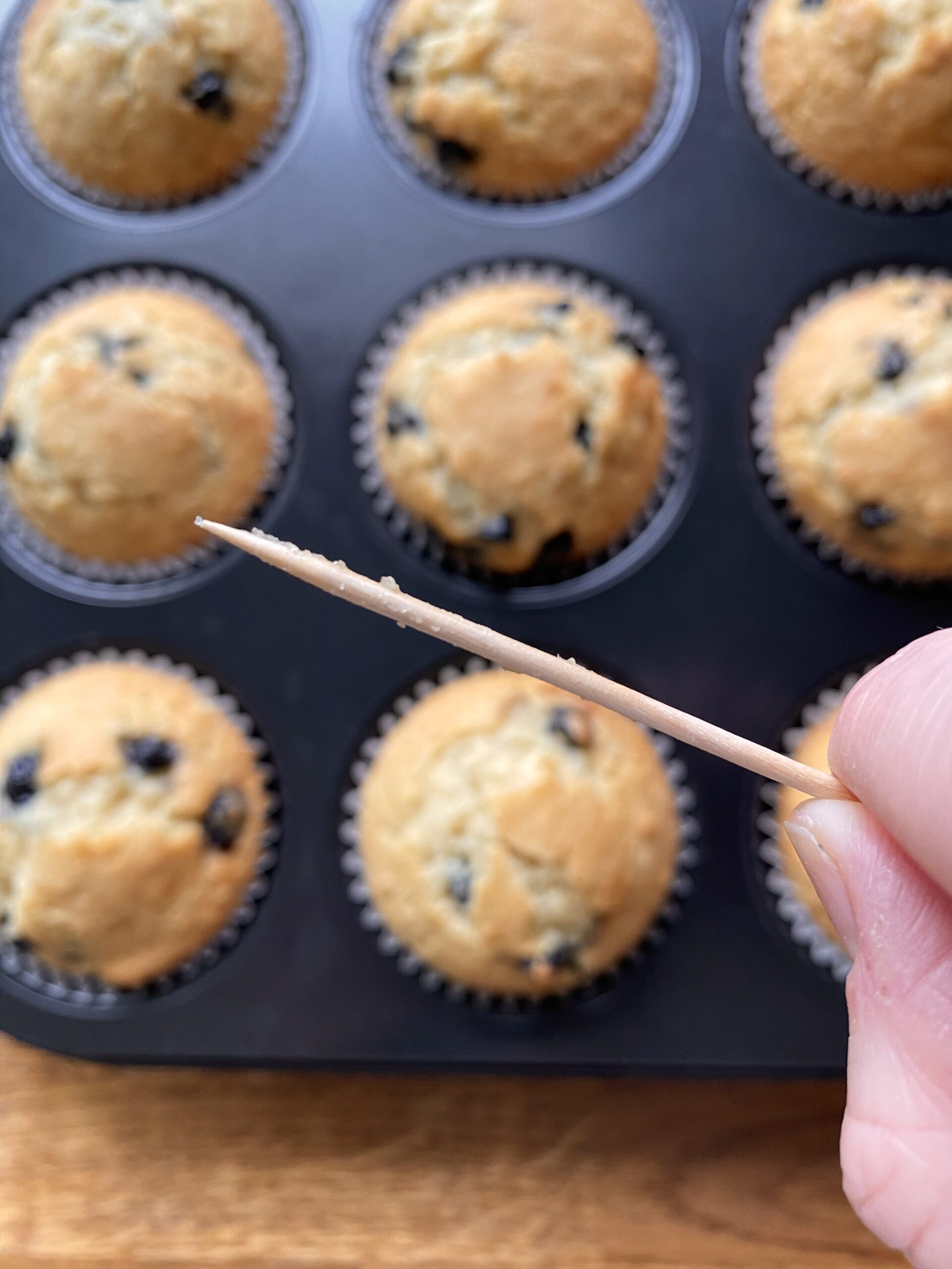 Close up of toothpick with just a few crumbs on it and no wet dough, with baked muffins in the background.